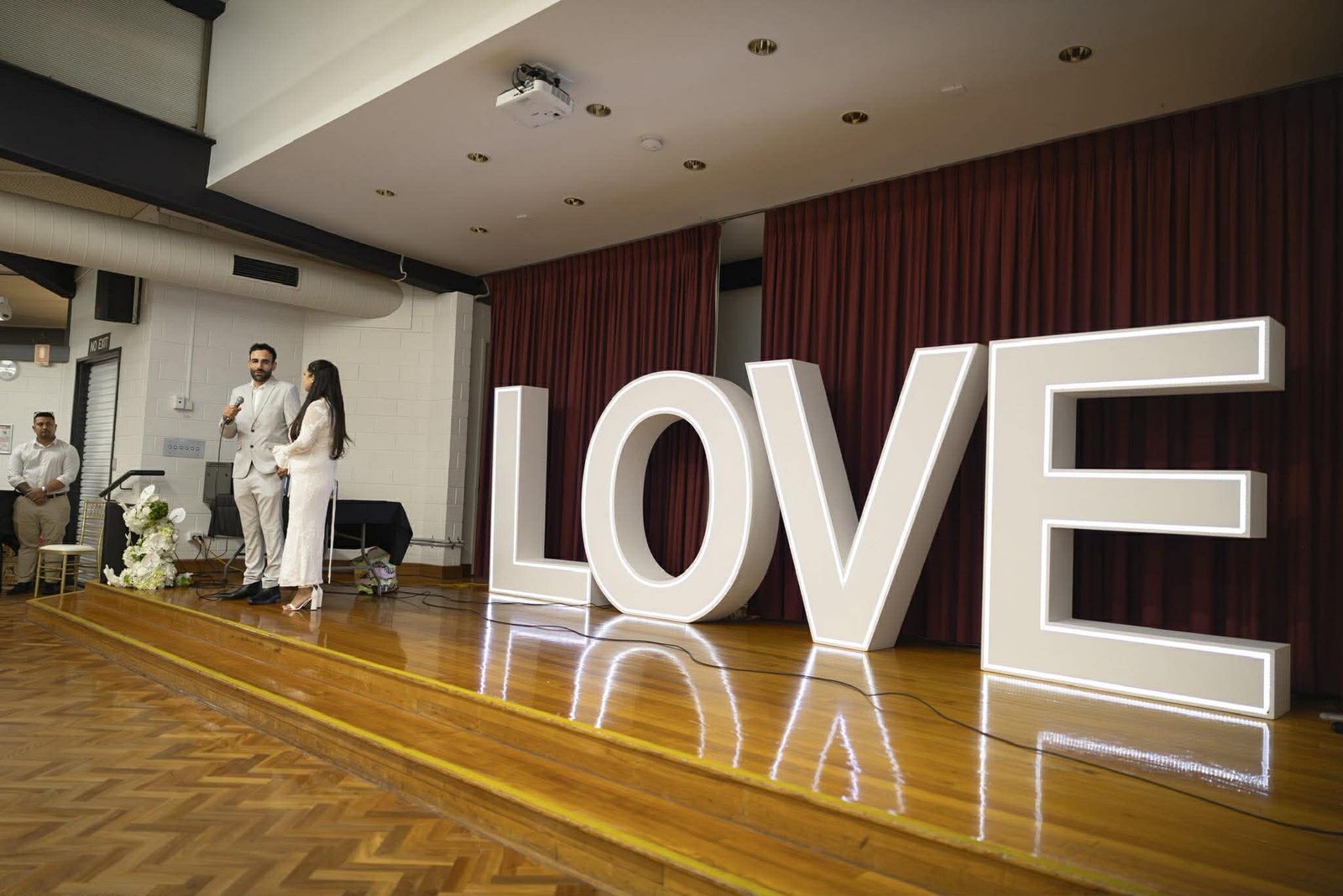 A couple speaking at a microphone beside giant lit LOVE letters, the light reflecting off the parquet floor